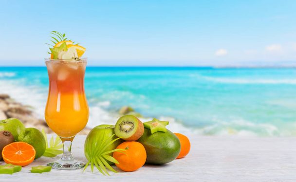 Zomer cocktail met diverse tropische vruchten rond. Appel, kiwi, sinaasappel, Mandarijn, mango, carambola's. Glas van Oranje drank met vervagen strand op achtergrond - Foto, afbeelding