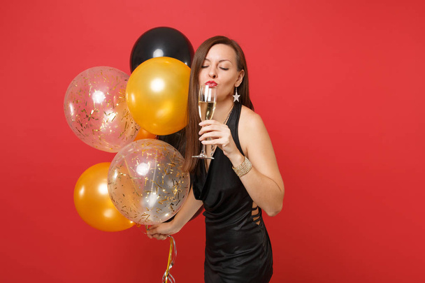 Bonita joven vestida de negro celebrando, bebiendo champán, sosteniendo globos aéreos aislados sobre fondo rojo. Día de San Valentín, Feliz Año Nuevo, concepto de fiesta de cumpleaños mockup
. - Foto, Imagen