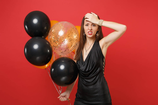 Tired young woman in little black dress suffering from headache putting hand on head holding air balloons celebrating isolated on red background. Happy New Year, birthday mockup holiday party concept. - Foto, immagini
