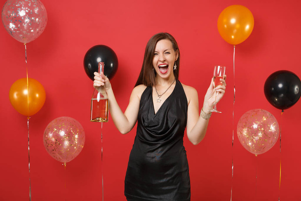 ガラス、明るい赤の背景の気球にシャンパンのボトルを保持している小さな黒いドレス記念に大喜びの若い女の子。バレンタインの日、幸せな新年、誕生日モックアップ休日党概念. - 写真・画像