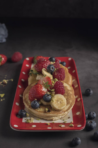 Pancakes with strawberries, raspberries, blueberries, banana, pistachios and syrup on a red plate with white dots. Vertical. Copyspace - Zdjęcie, obraz