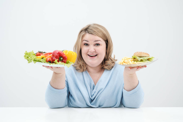 Young lush fat woman in casual blue clothes on a white background holding a vegetable salad and a plate of fast food, hamburger and fries. Diet and proper nutrition. - Photo, Image