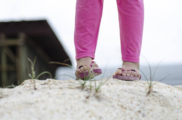 Pernas de criança close-up com sandália, em pé na areia sobre a profundidade rasa do campo
 - Foto, Imagem