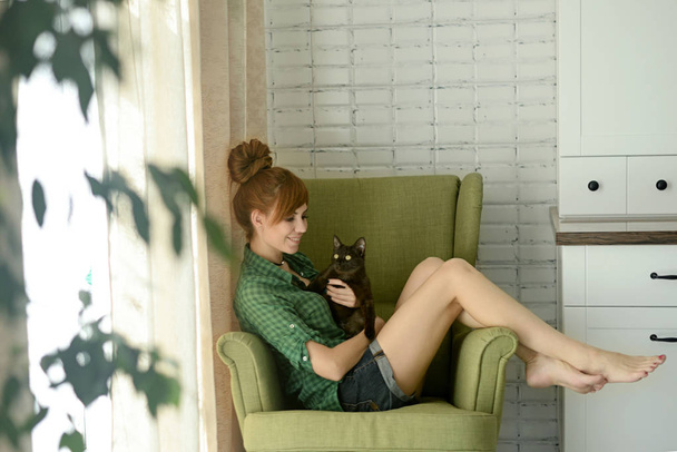 meisje in huis kleding, zittend in een stoel met een kat in haar armen - Foto, afbeelding