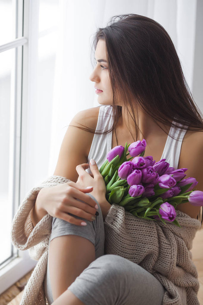 Attractive young woman with flowers indoors in the bedroom. Portrait of beautiful lady at home. Close up shot of female with tulips. - Foto, immagini