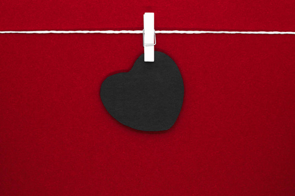 Μαύρο χαρτί καρδιά με χώρο αντίγραφο κρέμεται από λευκό σχοινί και συνδέονται με καρφίτσες πλυντηρίου λευκό σε κόκκινο φόντο - Φωτογραφία, εικόνα