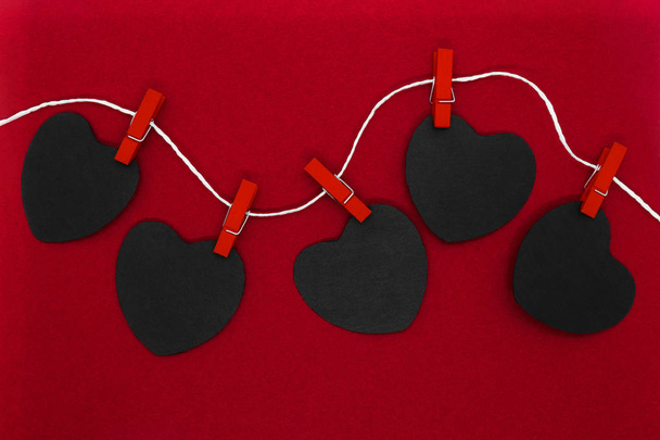 Black paper hearts with copy space hanging on white rope and attached with red laundry pins on red background - Photo, Image
