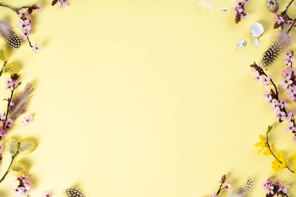 Easter holiday background. Flat-lay of tender Spring cherry blossom flowers on branches, feathers, quail eggs over pastel yellow background, top view - Photo, image