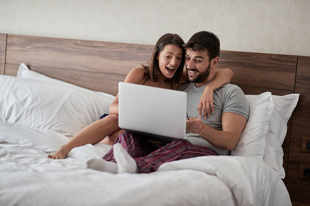 Happy young couple lying on a bed with computer - Beautiful married couple watching sex video on laptop laughing together - People, sexual, technology concept - Foto, imagen
