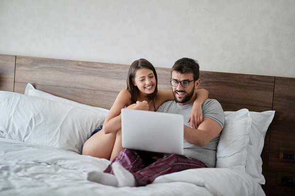 Young couple in the bed with laptop and surfing on internet. Modern lifestyle concept - Fotografie, Obrázek