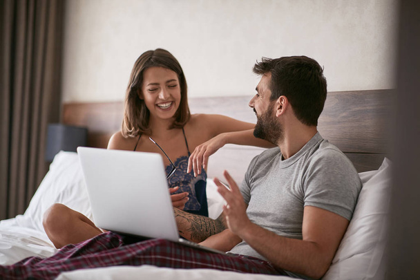 Young smiling couple discussion in the bed with laptop and surfing on internet. - Photo, Image
