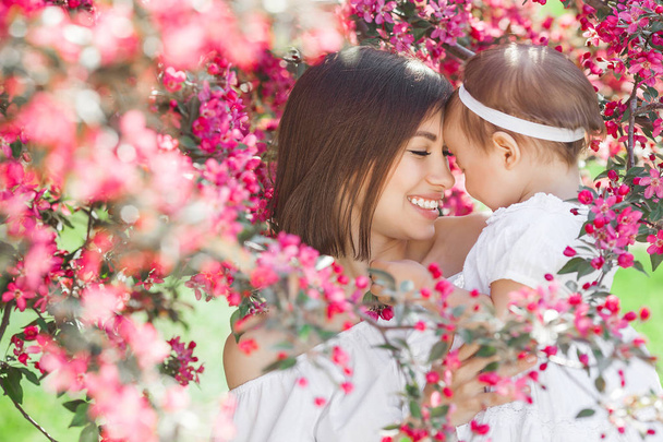 Portrait of young beautiful mother with her little baby girl. Close up still of loving family. Attractive woman holding her child in pink flowers and smiling. - Photo, Image