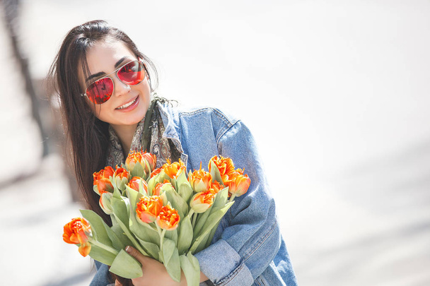 Mujer atractiva joven sosteniendo flores al aire libre. Mujer urbana vistiendo ropa casual sobre fondo urbano. Hermosa mujeren la ciudad
. - Foto, Imagen