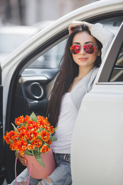 Молодая привлекательная женщина в машине с коробкой подарков, подарками и цветами. Прекрасная дама в весеннее время с букетом тюльпанов. Женщина в автомобиле
. - Фото, изображение