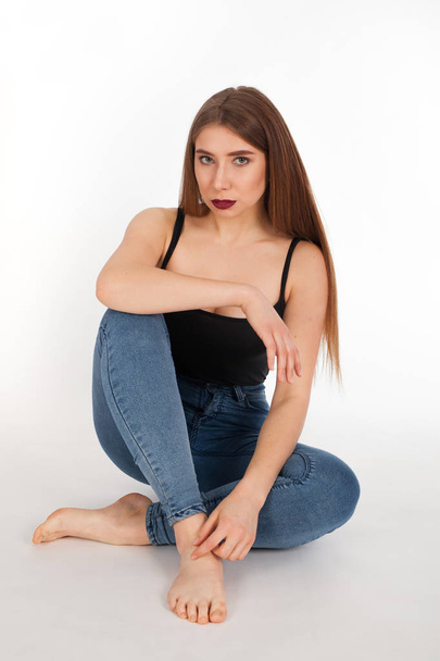 Portrait of a young beautiful girl with make-up isolated on white background. A woman in jeans and a t-shirt is sitting and looking at the camera. - Foto, Bild