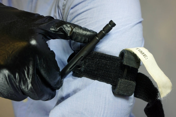 Άτομο σε μαύρο ιατρικά γάντια ισχύει το τουρνικέ για το χέρι του να αποτρέψει την αιμορραγία κατά τη διάρκεια των πρώτων βοηθειών. Ασθενή τραύμα. Την καταπολέμηση της τακτικής του εξοπλισμού. Εφαρμογή καταπολέμηση τουρνικέ - Φωτογραφία, εικόνα