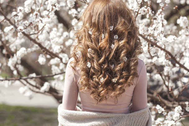 jong krullend meisje staan haar rug naar de camera op de bloemen achtergrond met bloemen in haar haar - Foto, afbeelding