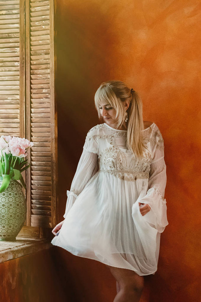 Jeune femme blonde en robe blanche dansant par la fenêtre, bouquet de fleurs roses fraîches dans un beau vase, mur de sienne lumineux en toile de fond. Romantique, amour, concept de bonheur
 - Photo, image
