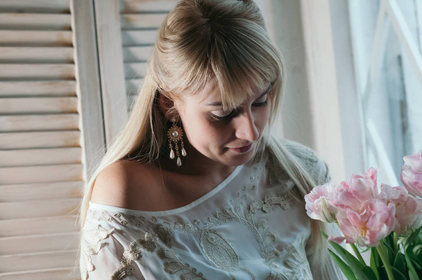 Une jeune femme blonde tenant un bouquet de fleurs roses fraîches près de la fenêtre, doucement souriante, image de mise au point douce. Romantique, amour, concept de bonheur
 - Photo, image