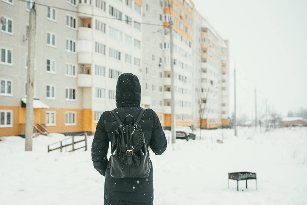 a back shot of a Beautiful youngwoman dressed in a black winter jacket with leather backpack walks in the snowy City - Fotografie, Obrázek