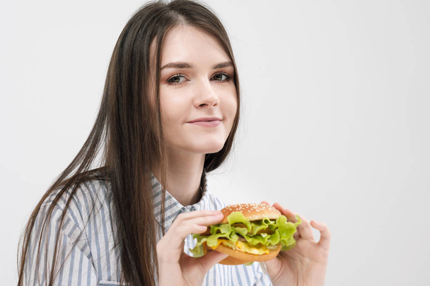 Slender brunette girl holds in her hands a hamburger. On a white background, the theme of diet and proper nutrition, choice and opposition. - Фото, изображение
