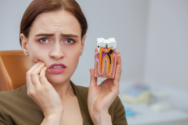 Young woman suffering from toothache, looking to the camera in despair, holding tooth mold showing cavity. Close up of a female patient with aching teeth visiting dental clinic. Dentistry, oral care - Photo, Image