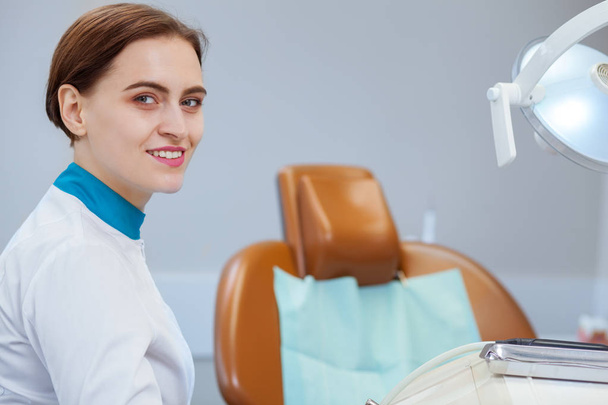 Attractive female doctor smiling to the camera over her shoulder. Professional dentist waiting for her patient at the dental clinic, smiling joyfully, copy space. Feminism, success, women concept - Photo, Image