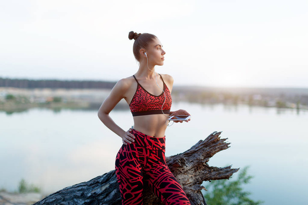 A beautiful Caucasian girl in bright sportswear and leggings rests after a run with the phone in her hands and headphones, listening to music. Sportswoman stands on the background of beautiful nature - Fotoğraf, Görsel