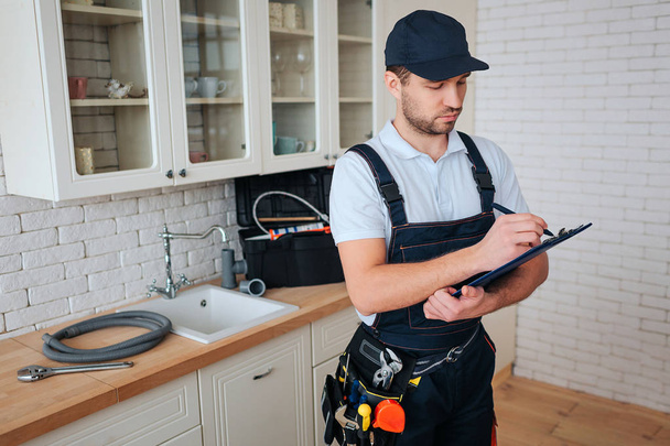 Busy young handyman stan in kitchen and white on plastic tablet. Toolbox and hose on desk. Daylight. Man in uniform. - Photo, image
