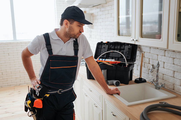 Thoughtful busy handyman stand in kitchen at sink and look at it. Toolbox and hose on desk. Guy wear uniform and belt with tools. - 写真・画像