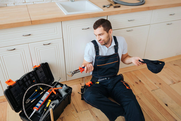 Confused young man sit on floor in kitchen and look at toolbox. He hold wrench. Tired man has rest. - Фото, изображение