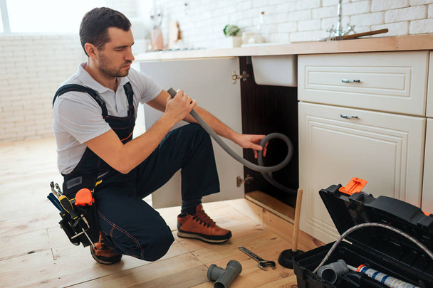 Concentrated young worker doing repairing in kitchen. He sit in squad pose and hold hose. Piece of pipe on floor. Opened toolbox. - Foto, imagen