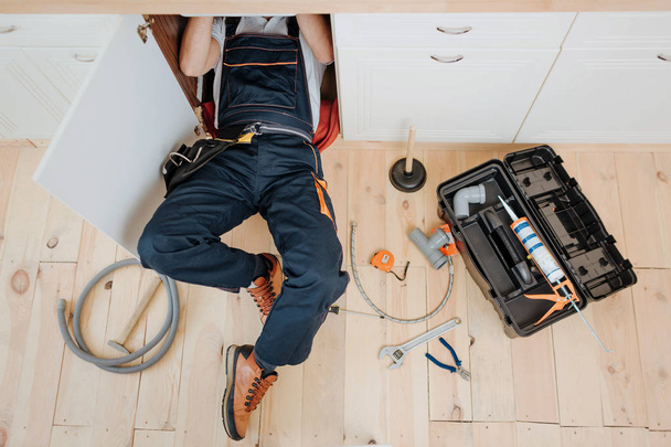 Mann in Uniform arbeitet in Küche unter Spüle. er repariert undichtes Wasser. Schlauch und Werkzeug auf dem Boden. Werkzeugkiste geöffnet. - Foto, Bild