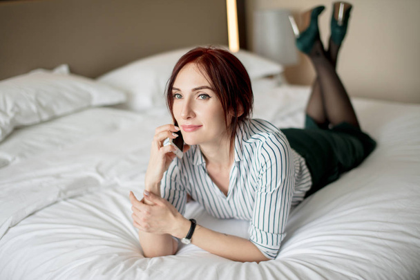 Elegant stylish businesswoman wearing high-heeled shoes lying on bed in hotel - Photo, Image