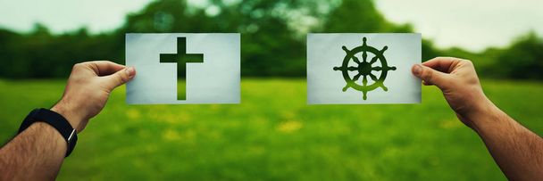 Religion conflicts as global issue concept. Two hands holding different faith symbols, Buddhism vs Christianity belief over green field nature. Relations between different people doctrines and cult. - Photo, Image