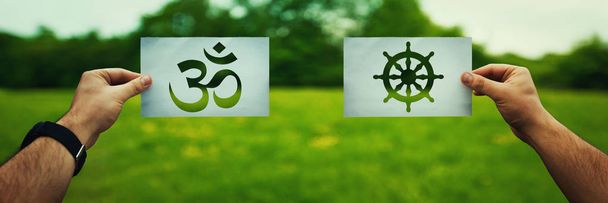 Religion conflicts as global issue concept. Two hands holding different faith symbols, Buddhism vs Hinduism belief over green field nature. Relations between different people doctrines and cult. - Photo, Image