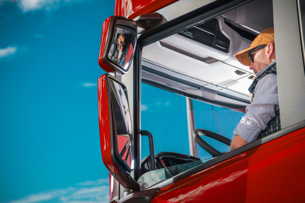 Θέμα βιομηχανία μεταφορών. Καυκάσιος φορτηγό οδήγησης στα 30 του στο εσωτερικό της καμπίνας κόκκινο ημι.  - Φωτογραφία, εικόνα