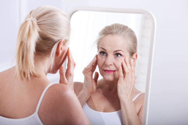 Middle aged woman looking at wrinkles in mirror. Plastic surgery and collagen injections. Makeup. Macro face. Selective focus - 写真・画像