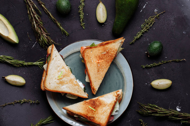 Three delicious fresh sandwiches in a plate on a black background with green vegetables, fruits and herbs on a black background. Top view, closeup. Cut avocado and feijoa, rosemary and cumin. - Фото, изображение