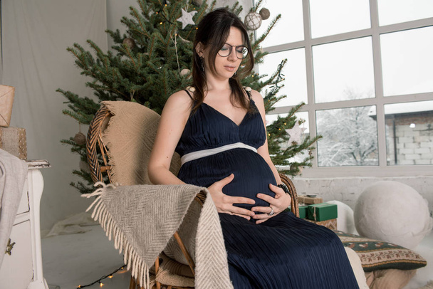 joven chica embarazada bonita en gafas como un triángulo para las mujeres embarazadas y sentarse en una silla cerca de un abeto
 - Foto, Imagen