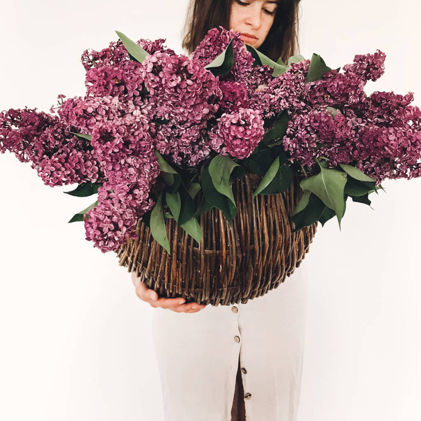 Hipster Girl в сільській лляній сукні, що тримає великі фіолетові бузкові квіти в плетеному кошику на білому тлі. Привіт, весна. Щасливий день матері. Букет з бузку в руках жінки. Міжнародний жіночий день
 - Фото, зображення