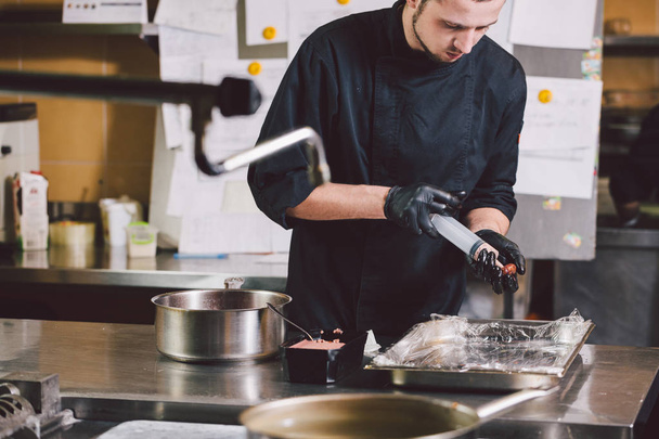 Thema koken. Een jonge blanke man in zwarte uniforme en latex handschoenen in een restaurant in de keuken. Koken vleesmolens in een BES met een spuit. Een schotel vijgen, of vijgen fruit vijgenboom Ficus Carian. - Foto, afbeelding
