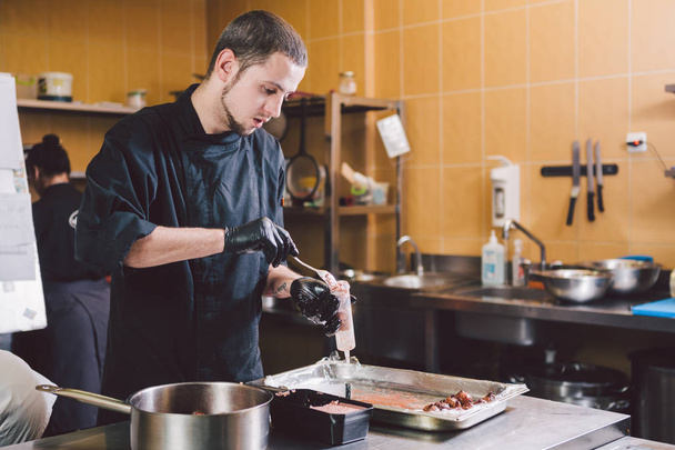 Theme cooking. A young Caucasian man in black uniform and latex gloves at a restaurant in the kitchen. Cooking mincers into a berry with a syringe. A dish of figs, fig fruit fig tree or Ficus Carian. - Fotografie, Obrázek