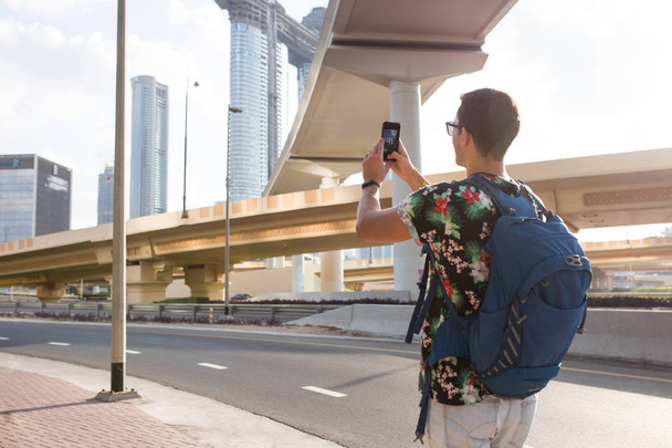 Joven turista atractivo en camiseta con mochila de atrás tomando fotos en el teléfono celular caminando soñadoramente por la ciudad moderna
 - Foto, Imagen