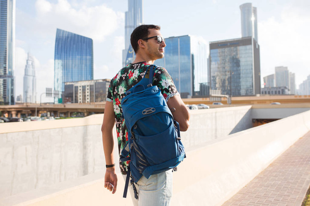 Mladý atraktivní muž v sluneční brýle a tričko s batohem zasněně dívá stranou chodit po městě s krásný mrakodrap na pozadí - Fotografie, Obrázek