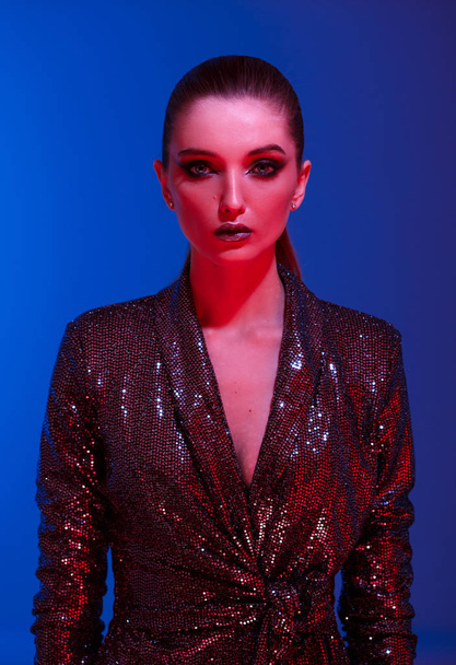 Trendy girl with long hair and stylish makeup in a black shining dress poses on the blue  background in neon light in the studio - Foto, afbeelding