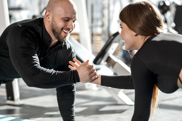 Brutal athletic man and young slender girl dressed in black sorts clothes are doing plank  holding hand in hand in the modern gym - 写真・画像