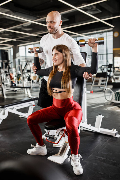 Beautiful slender girl in stylish sports clothes does a dumbbell bench press up sitting on a bench while the athletic man insures her in the modern gym - Fotografie, Obrázek