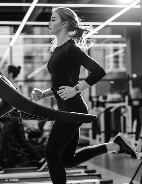 Black and white photo of the athletic girl dressed in a black sportswear running on the treadmill in the modern gym - Fotografie, Obrázek