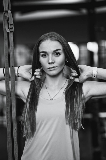 Black and white photo of the beautiful athletic girl dressed in sporty clothes standing in the gym with her hands on her hair - Фото, зображення
